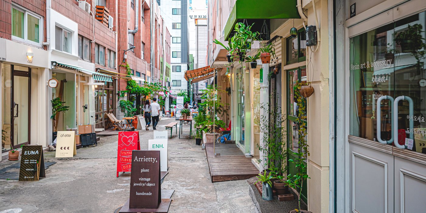 คนสองคนเดินในซอยถนนคาเฟ่จอนโพ ปูซาน สองข้างทางมีร้านเล็ก ๆ และกระถางต้นไม้