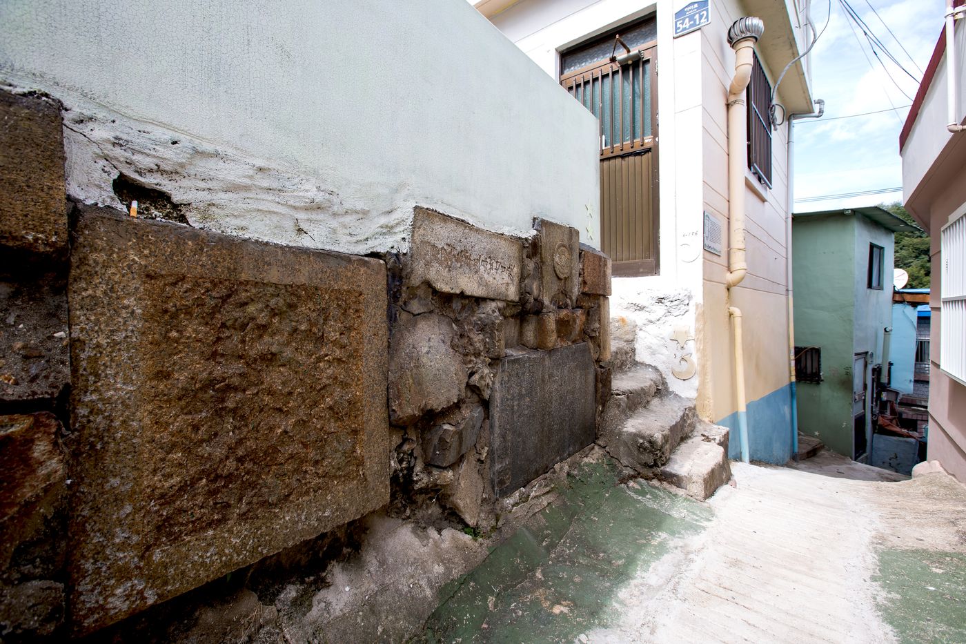 Steps and a wall built from old tombstones and altar stones, right beside a house number plate (54-12) — someone's actual home.