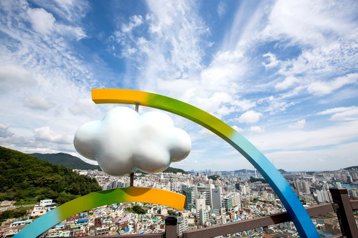 A rainbow-shaped sculpture with a cloud ornament at the viewpoint, with the Busan skyline beyond.