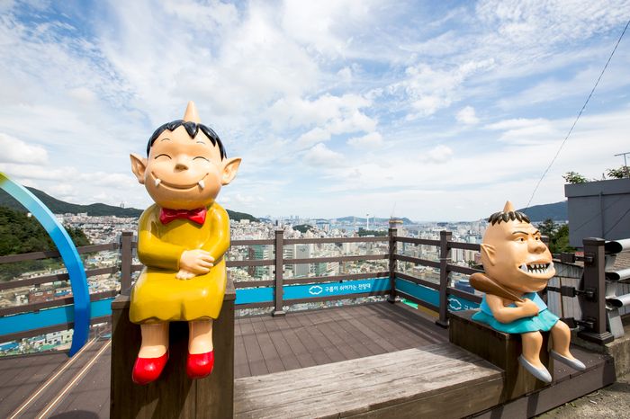 Two goblin-character statues sitting on the viewpoint railing, with the Busan skyline behind them.
