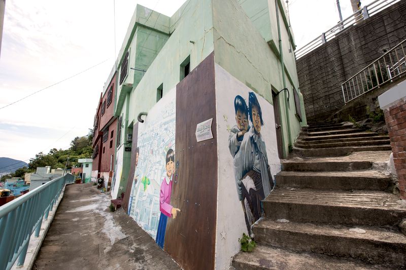 A mural on a wall beside the alley steps, showing a woman carrying a child on her back.